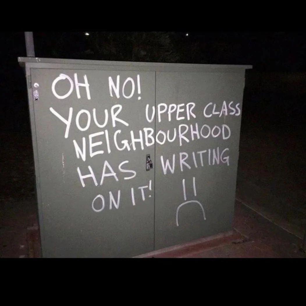 The meme is a photo of a street sign or a public wall with a message written in chalk or spray paint. The text reads: "In 2-3 sentences, describe this meme for someone who cannot see it."